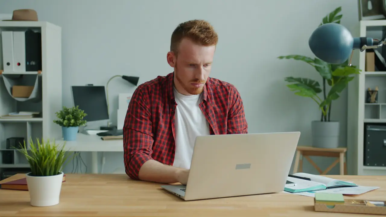 A man in a checked shirt working through paperwork on a laptop at a bright desk