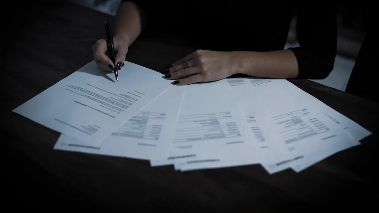 A hand signing one of several printed pages spread across a dark table