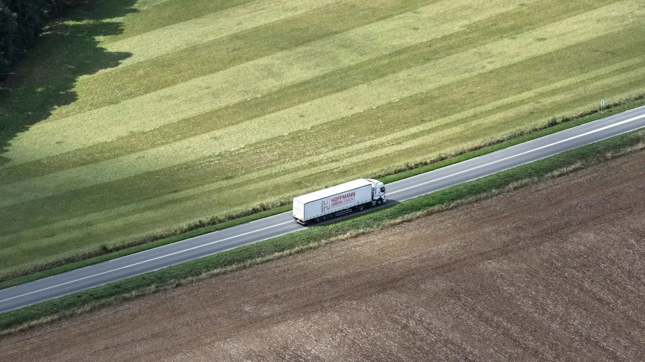 A white transport truck on a two lane highway between green and brown fields, seen from above