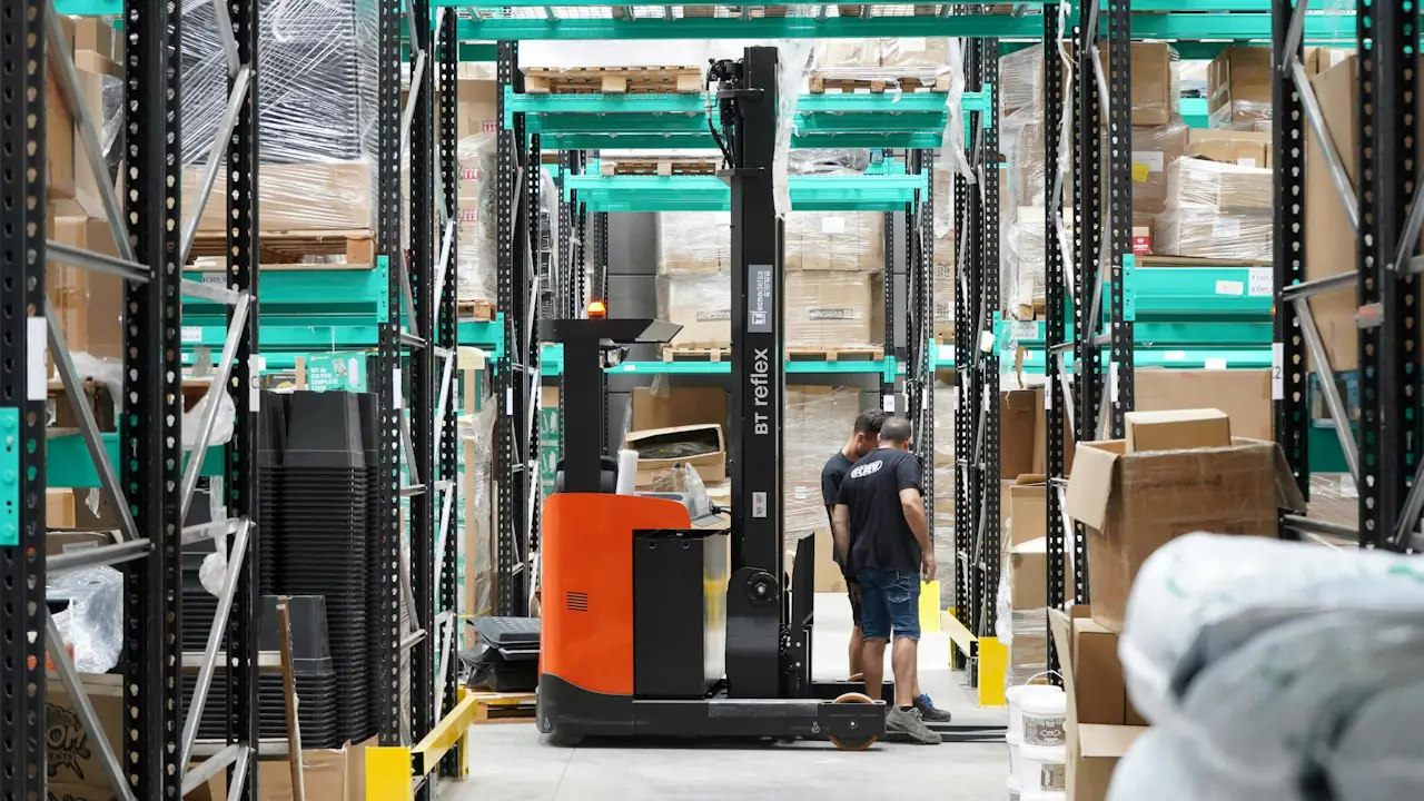 A worker beside an orange reach truck in a tall warehouse aisle stacked with boxes