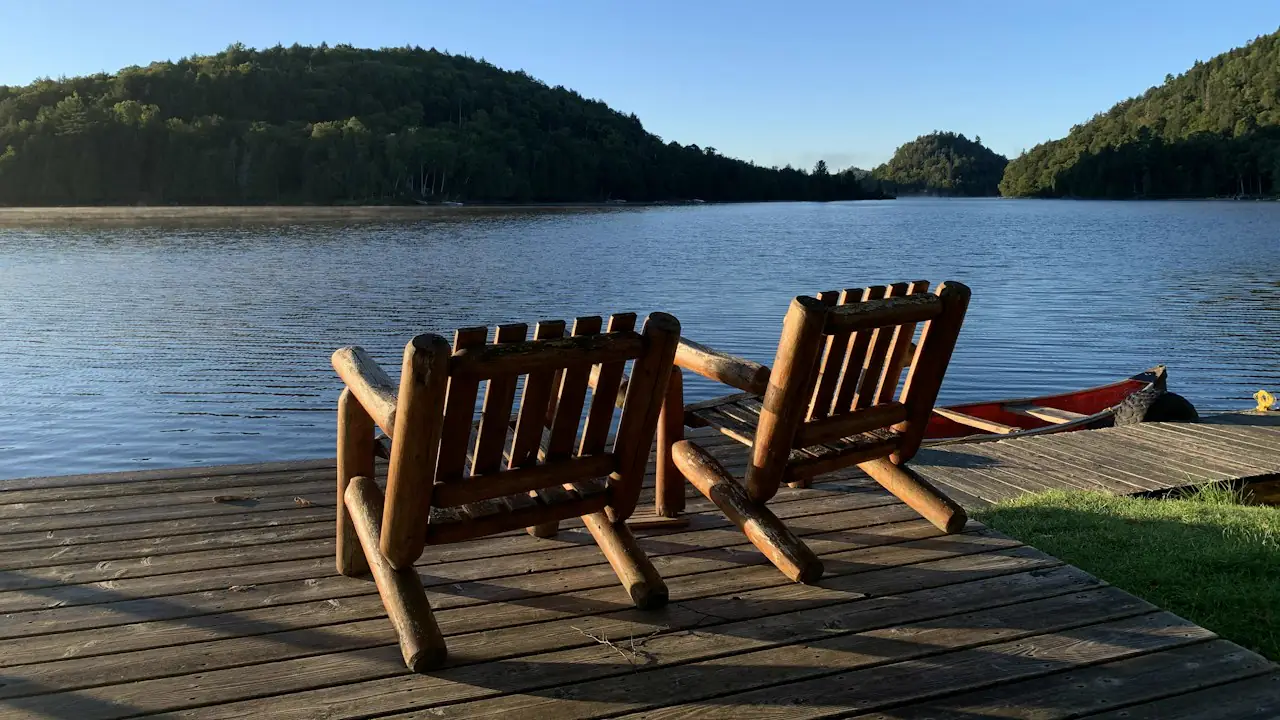 Two wooden chairs on a dock facing a calm lake and forested hills