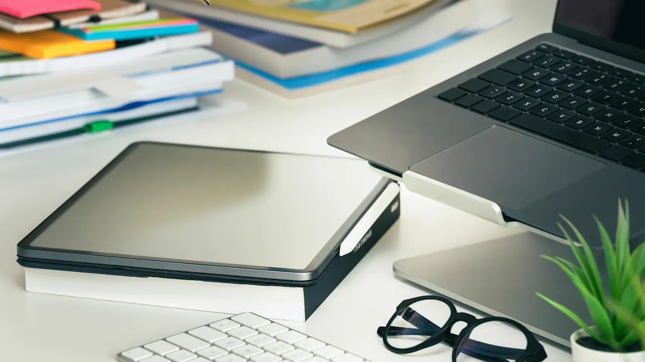 A laptop, a tablet, a keyboard and glasses on a white desk beside a small plant