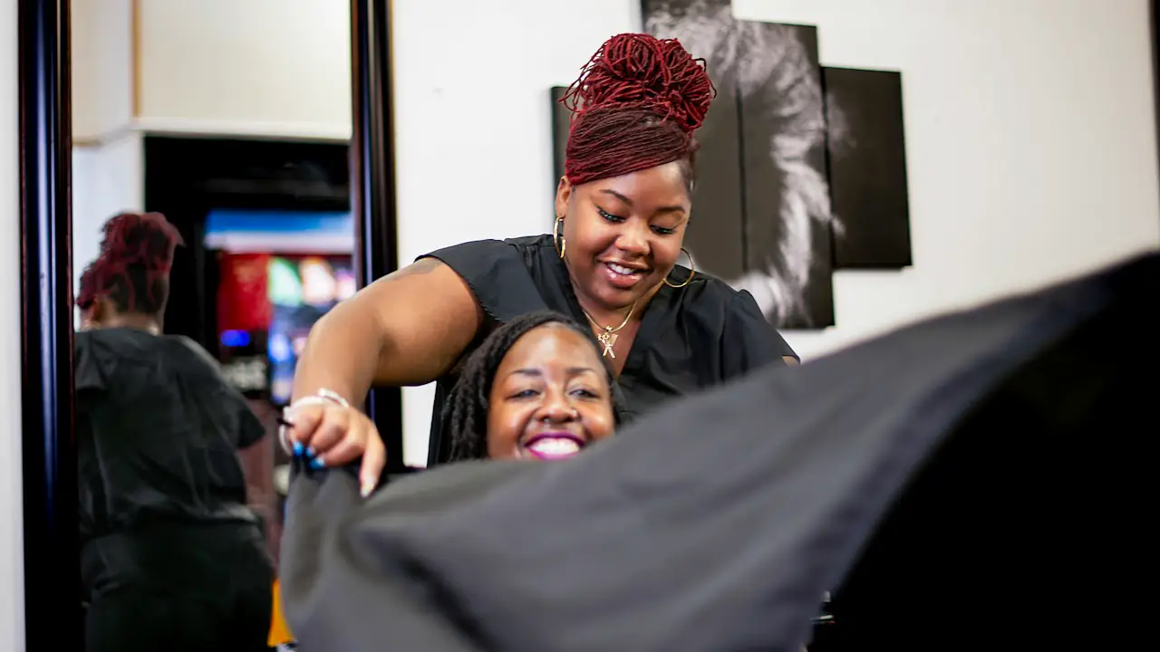 A smiling stylist draping a cape over a laughing client in a salon chair