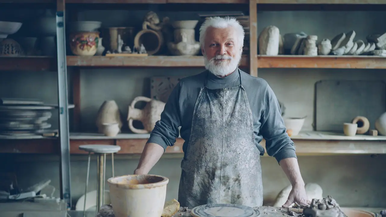 A smiling potter with a white beard standing at his wheel in a workshop full of pottery