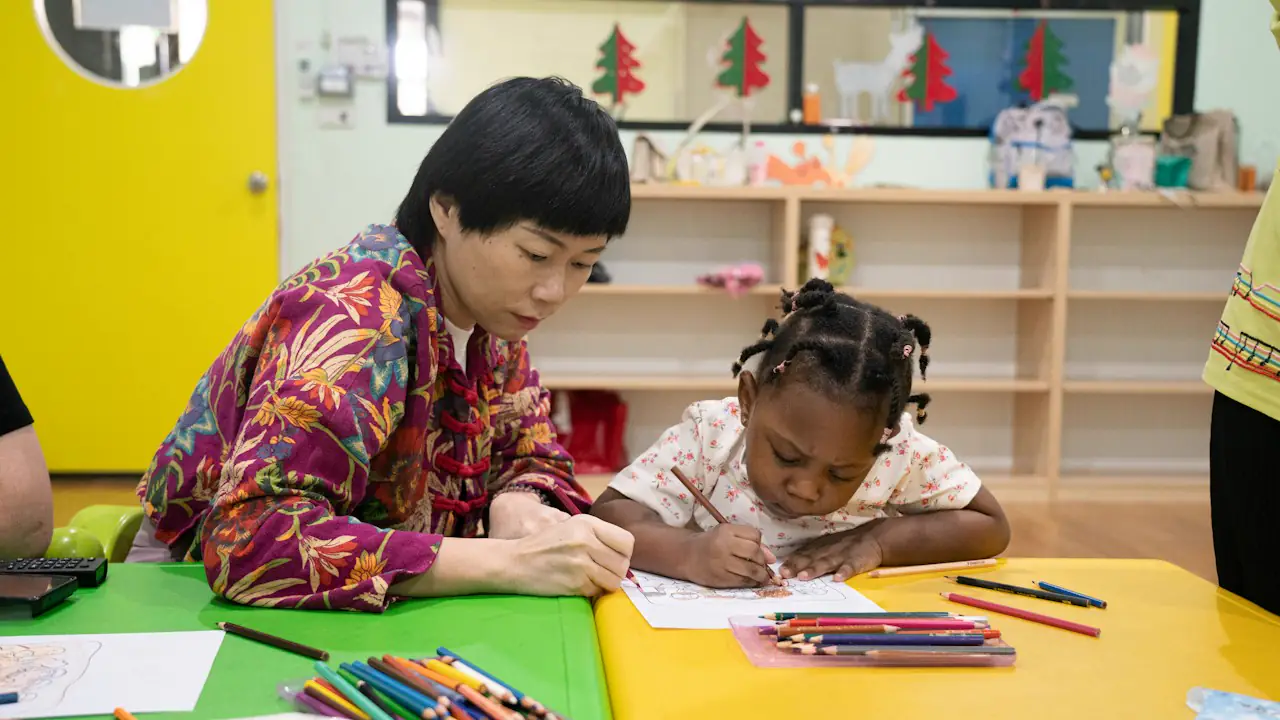 A child care worker drawing with a young girl at a bright classroom table
