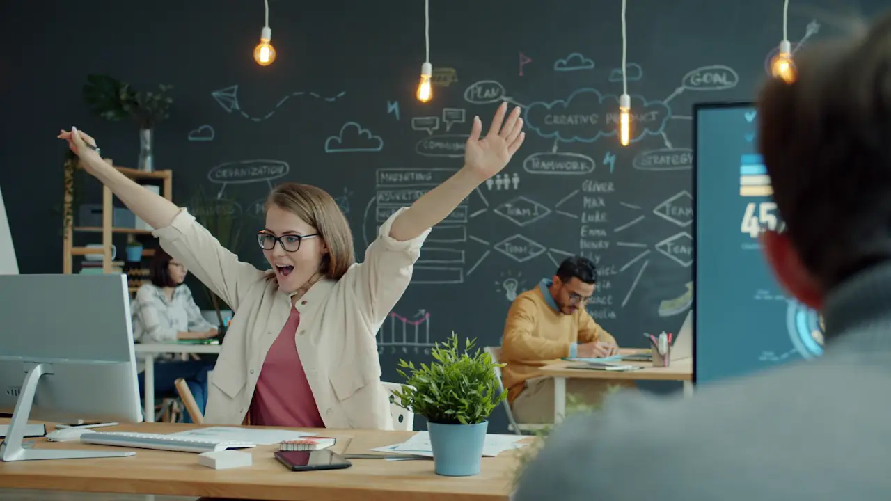 A woman throwing her arms up in celebration at her desk in a busy office