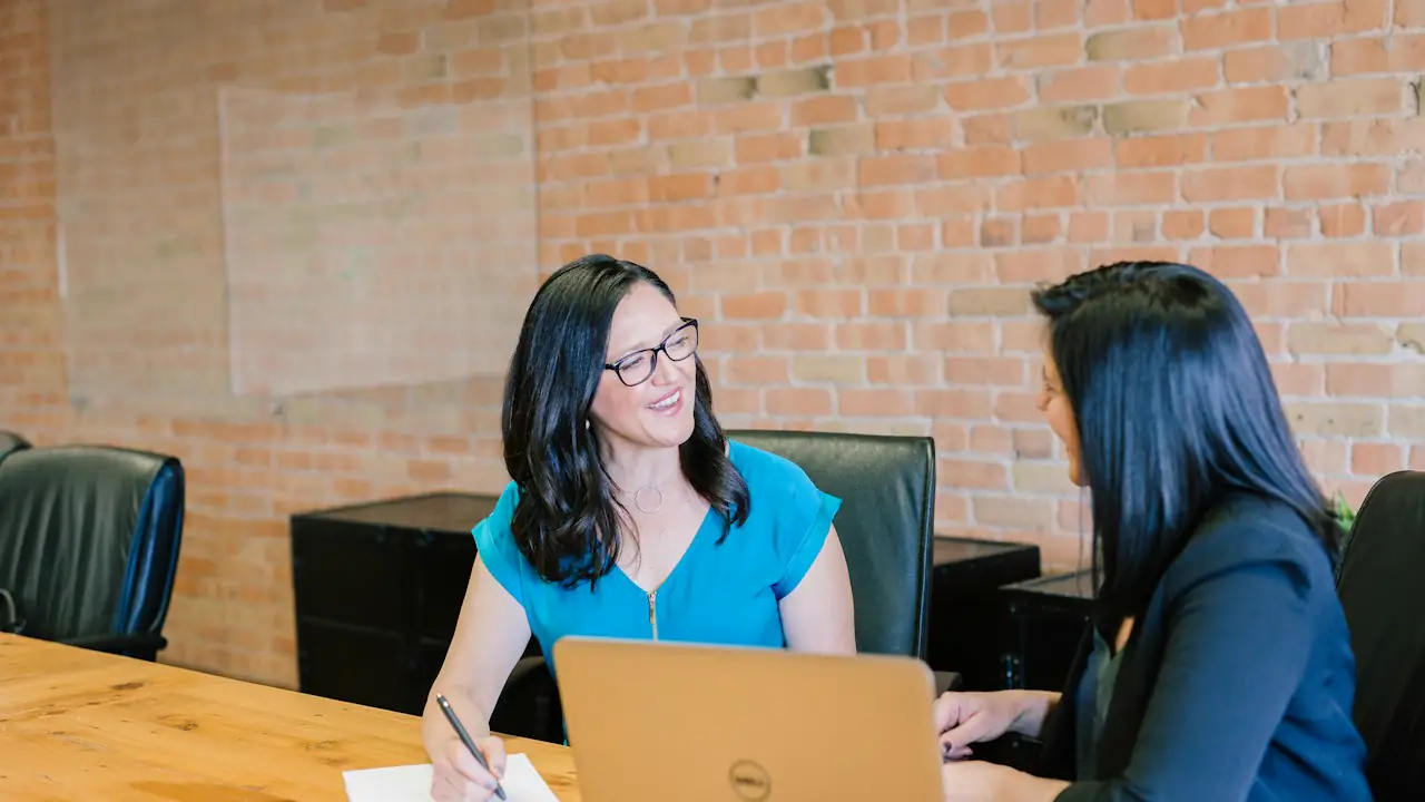 Two women talking over a laptop and notebook at a meeting table