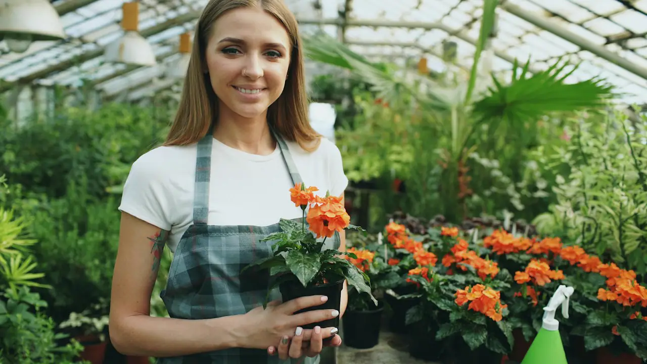 A smiling greenhouse owner holding a pot of orange flowers among her plants