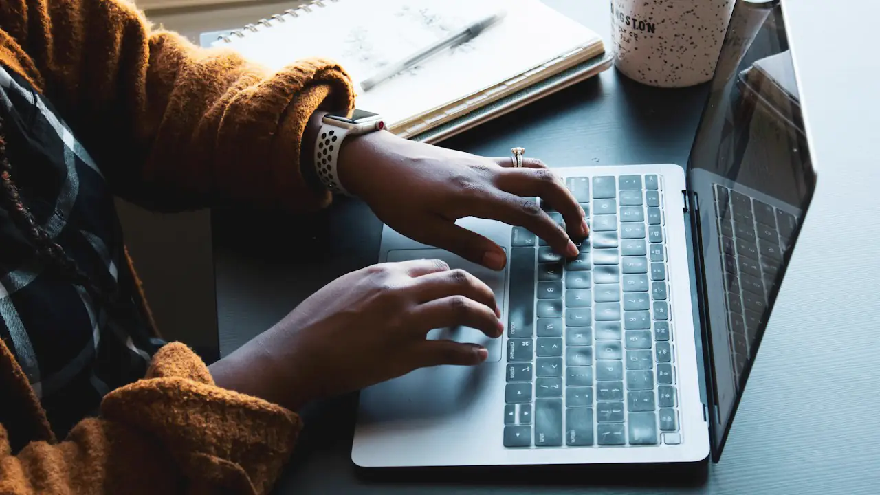 Hands typing on a laptop beside a notebook and a mug of tea
