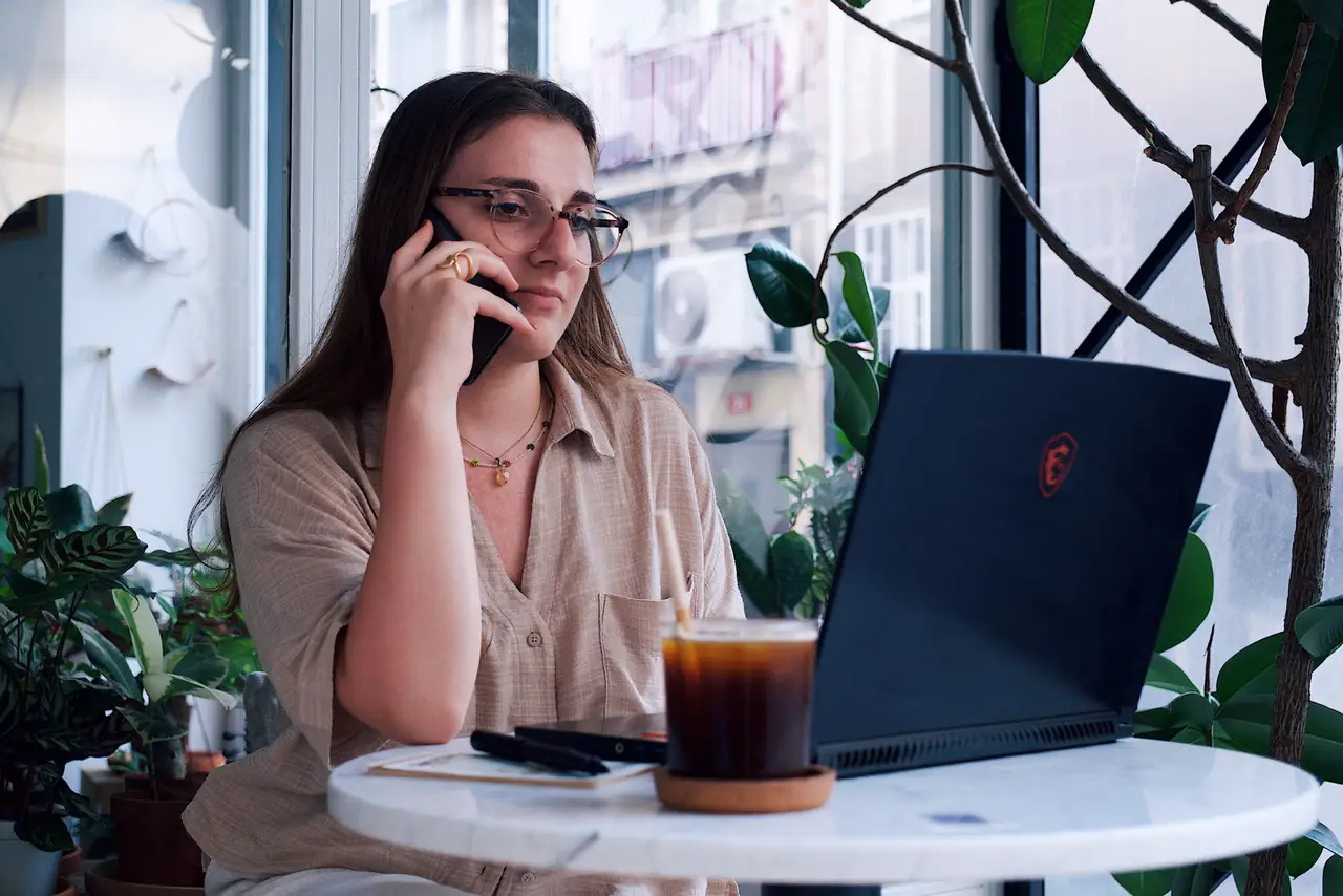 Consultant takes a phone call beside her laptop at a cafe table with plants