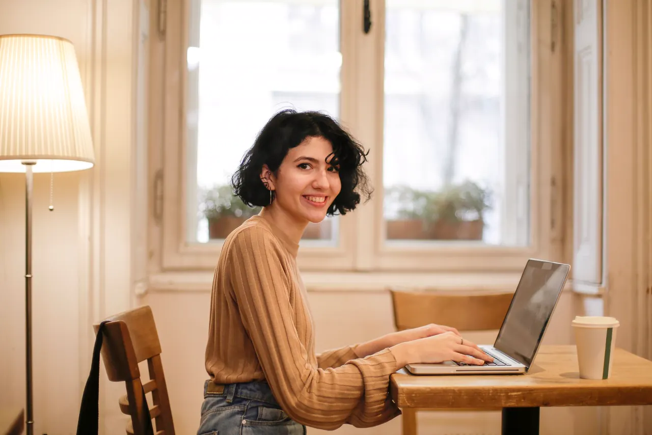 Smiling woman with short dark hair works on a laptop at a wooden table by a window