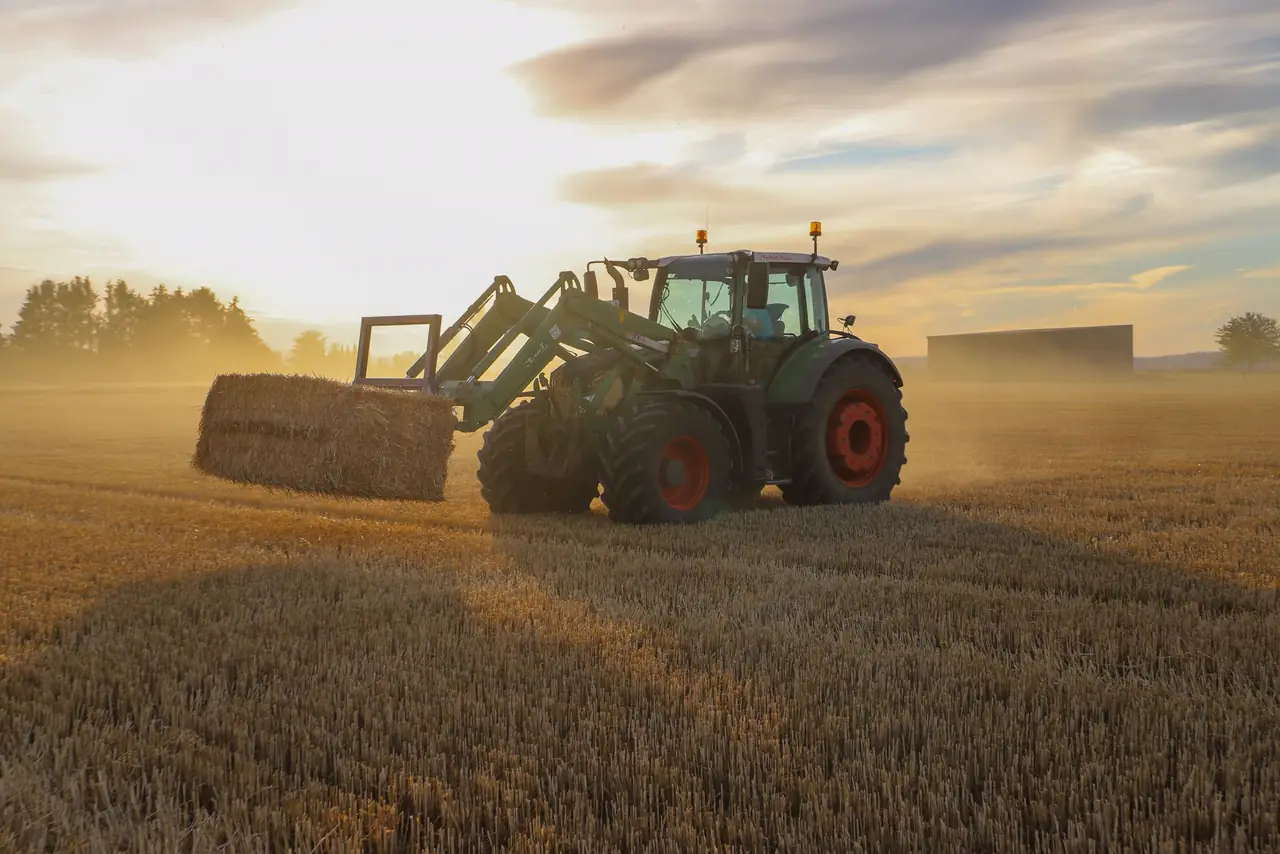Tractor lifts a hay bale in a field under a warm evening sky
