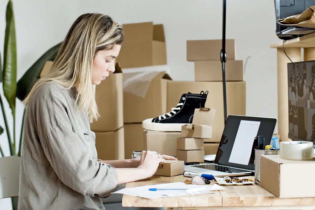 Seller packs boxes for her online store surrounded by packaging materials and a laptop