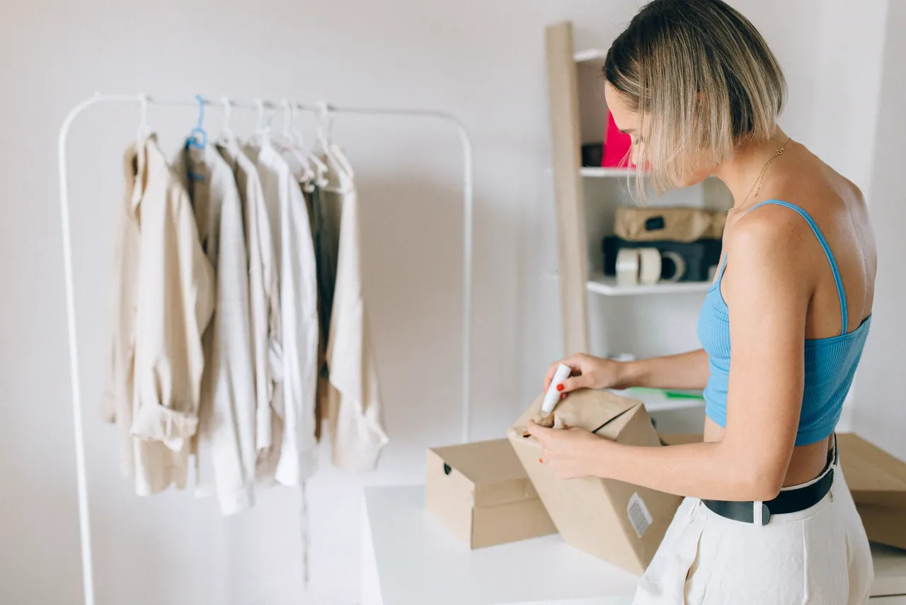 Woman packs a garment into a cardboard shipping box beside a clothing rail in a bright room