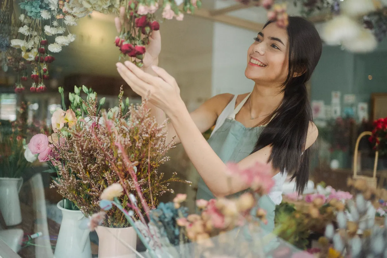 Smiling florist in an apron arranges dried and fresh flowers inside her colourful flower shop