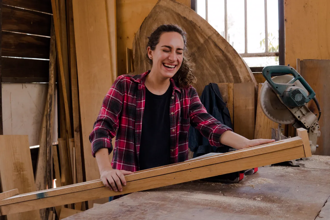 Laughing carpenter in a plaid shirt holds a long plank in a sunlit woodworking shop