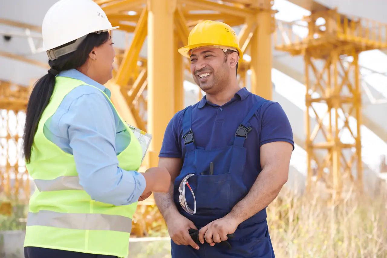 Two construction workers in hard hats smile and talk on a sunny building site