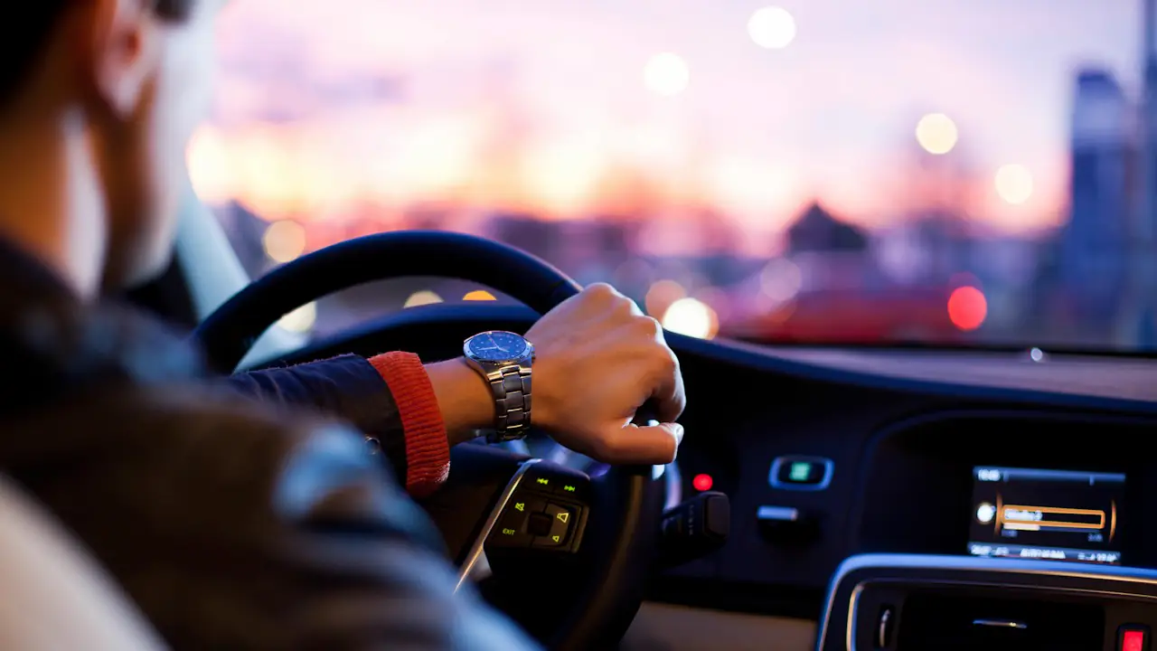 A driver's hand on the steering wheel with city lights glowing at dusk