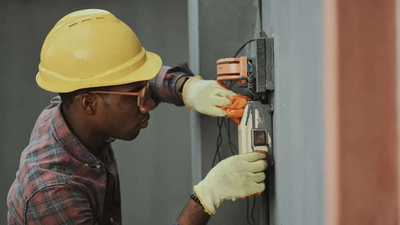An electrician in a yellow hard hat and gloves working on a wall panel