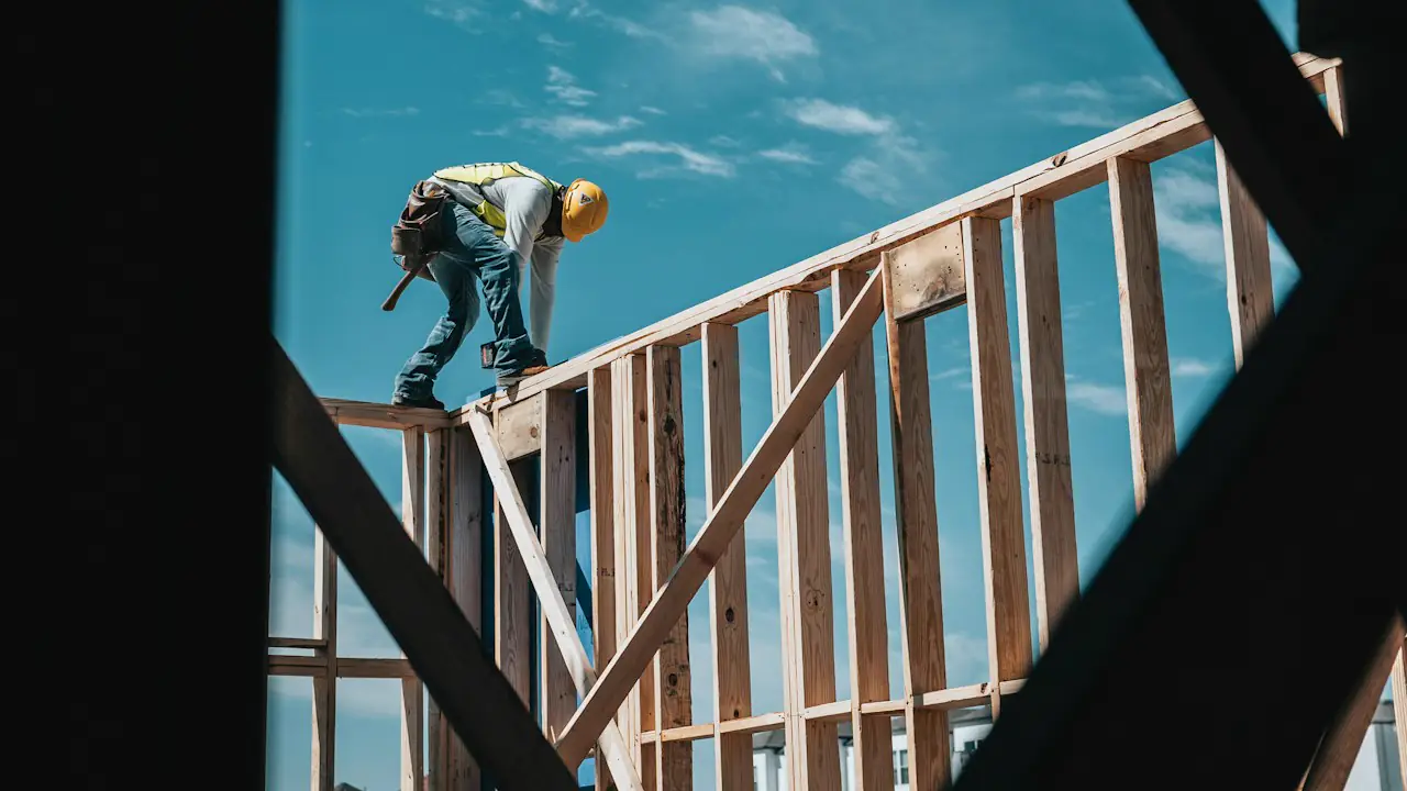 A carpenter in a hard hat working on top of a timber wall frame against a blue sky