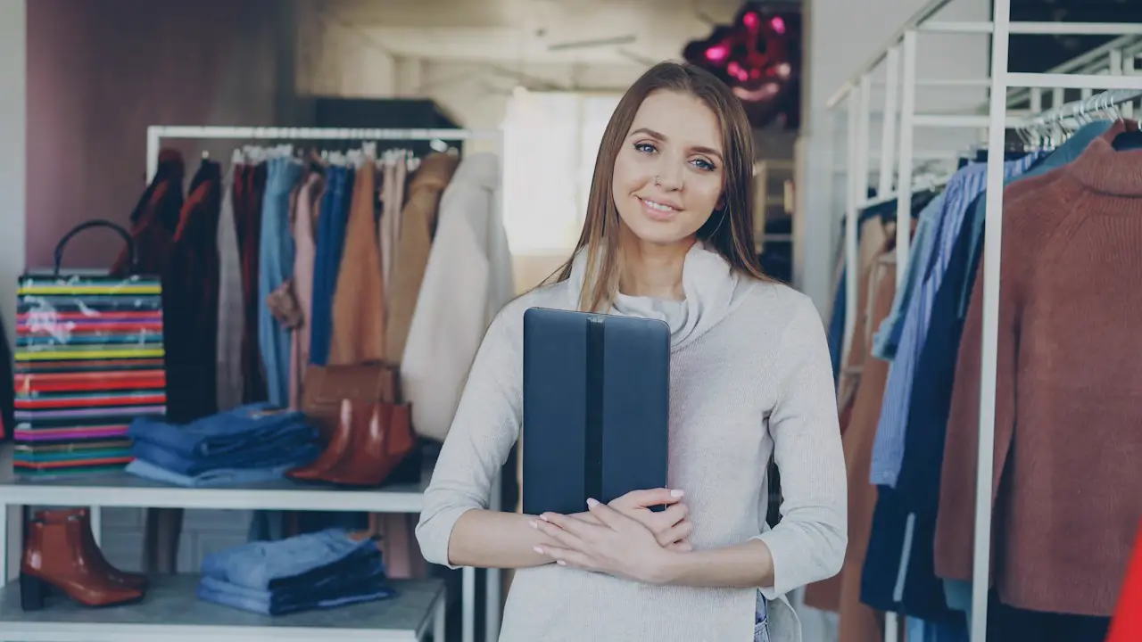 A smiling shop owner holding a tablet among racks of clothes in her boutique