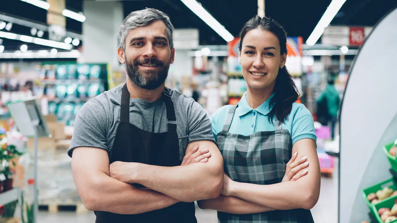 Two smiling grocery store employees in aprons standing in a produce aisle