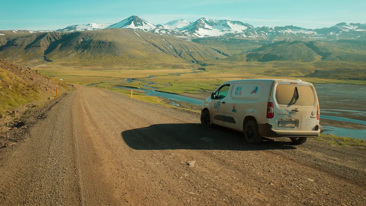 A white work van parked on a gravel road in a wide valley below snowy mountains