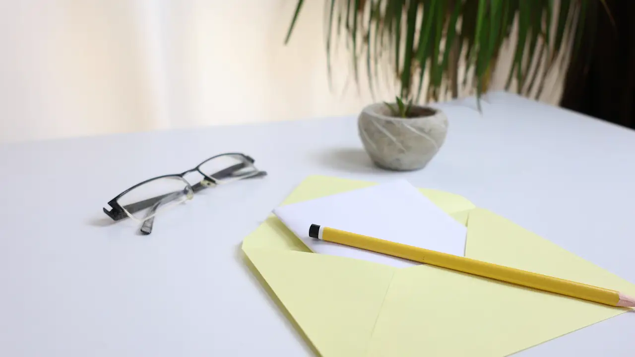An opened yellow envelope with a note, glasses and a pencil on a white desk