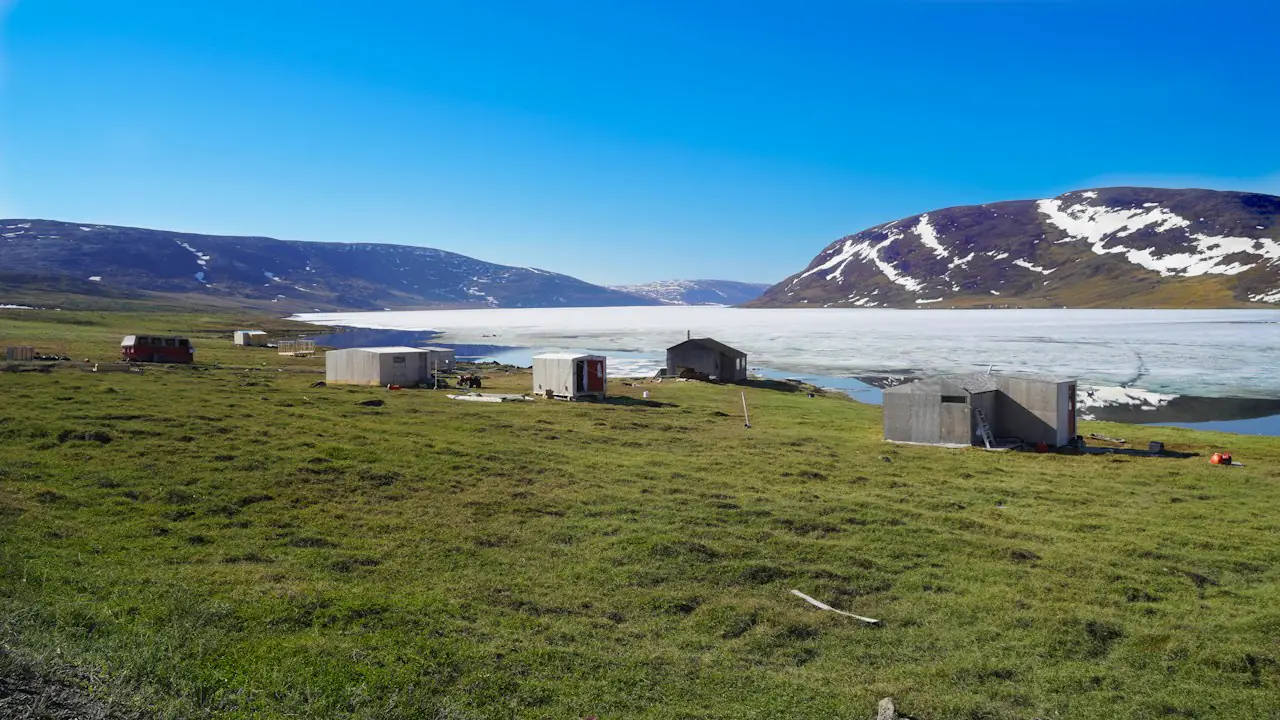 Cabins on green summer tundra beside sea ice, with snowy hills behind