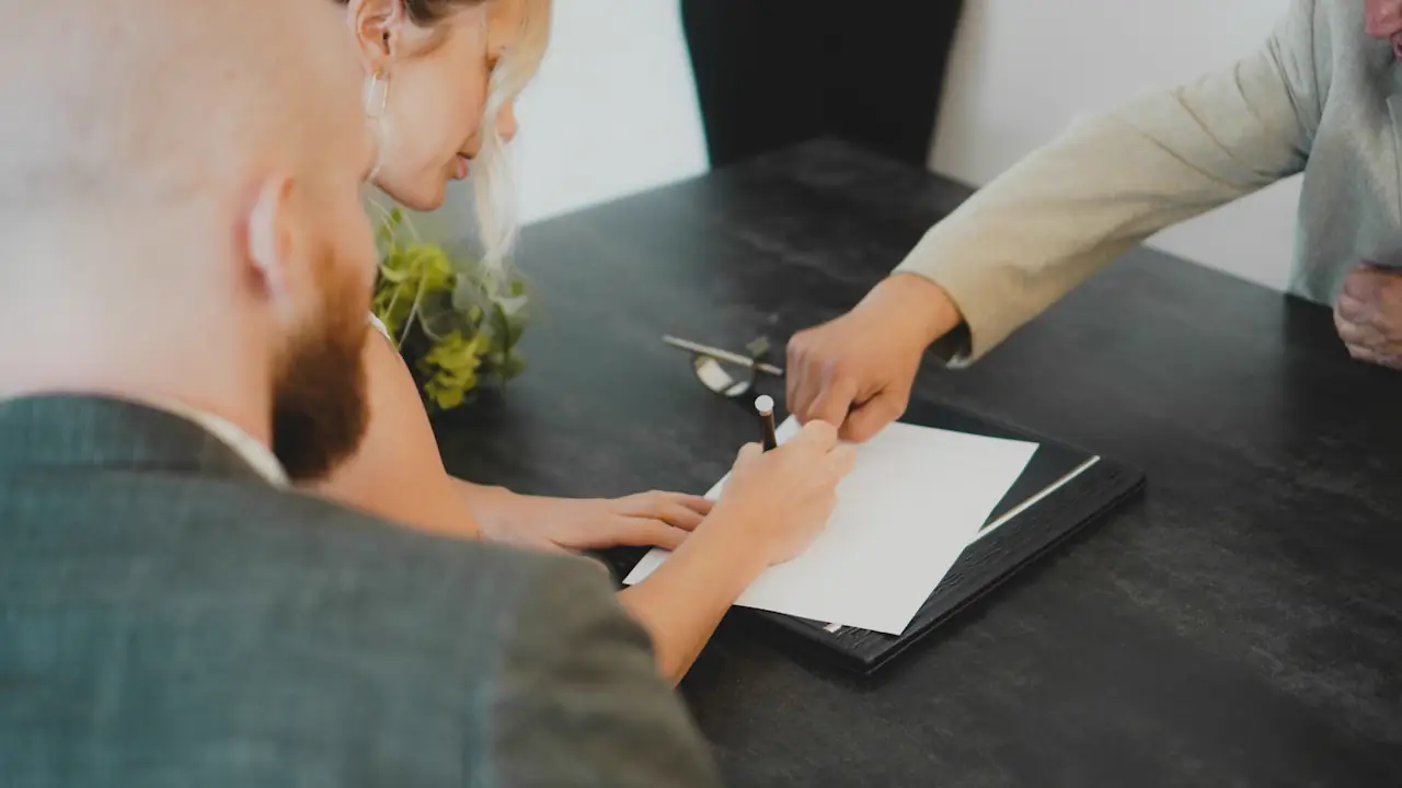 Two people signing a document on a clipboard across a dark table