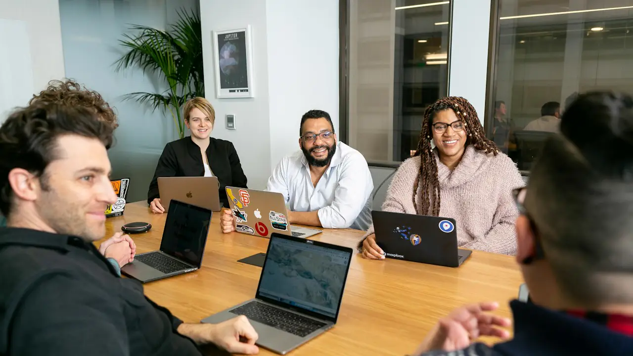 A small team laughing together around a meeting table with laptops