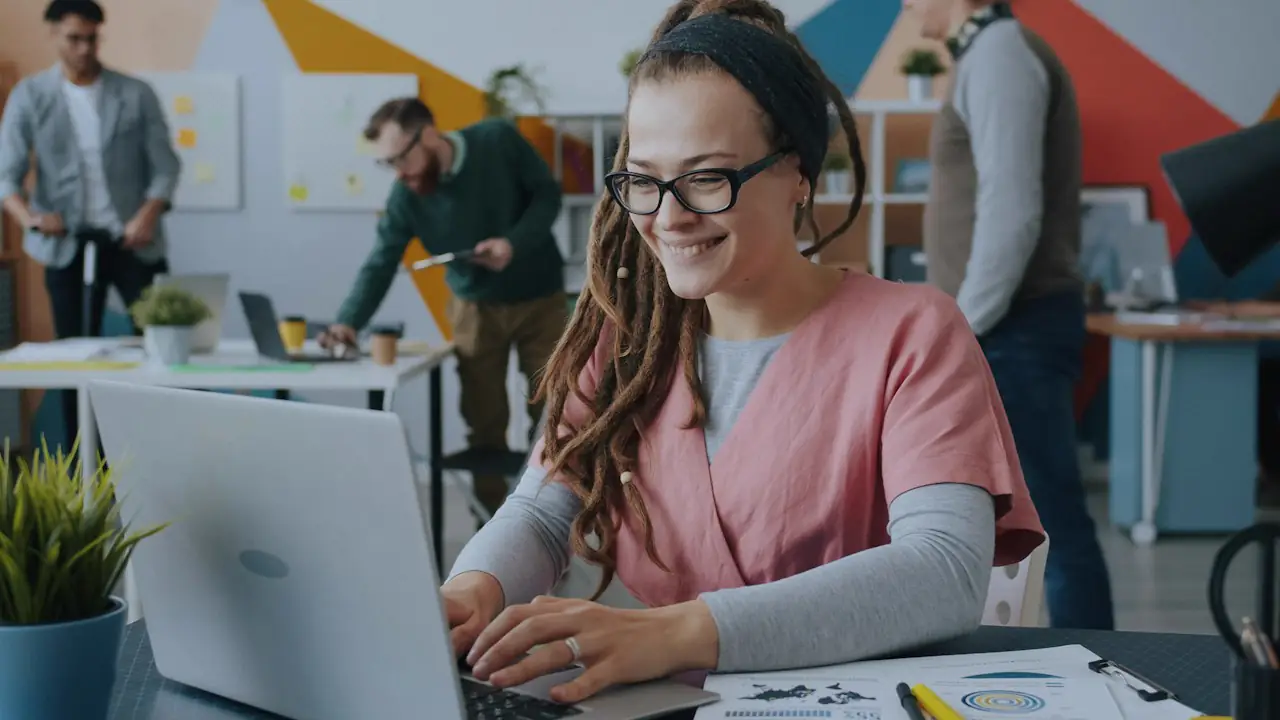A smiling woman in glasses working at a laptop in a busy open office