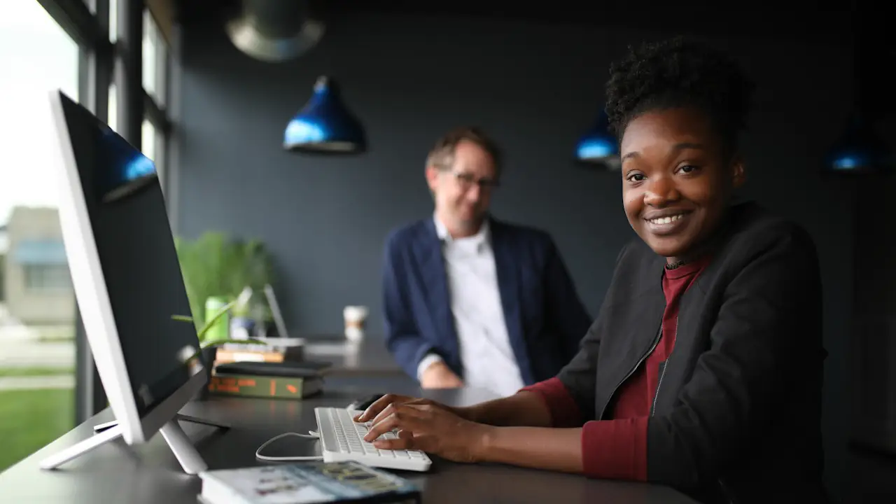 A smiling woman at her computer in a modern office, with a colleague working behind her