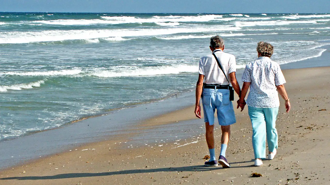 An older couple walking hand in hand along a sandy beach beside the surf