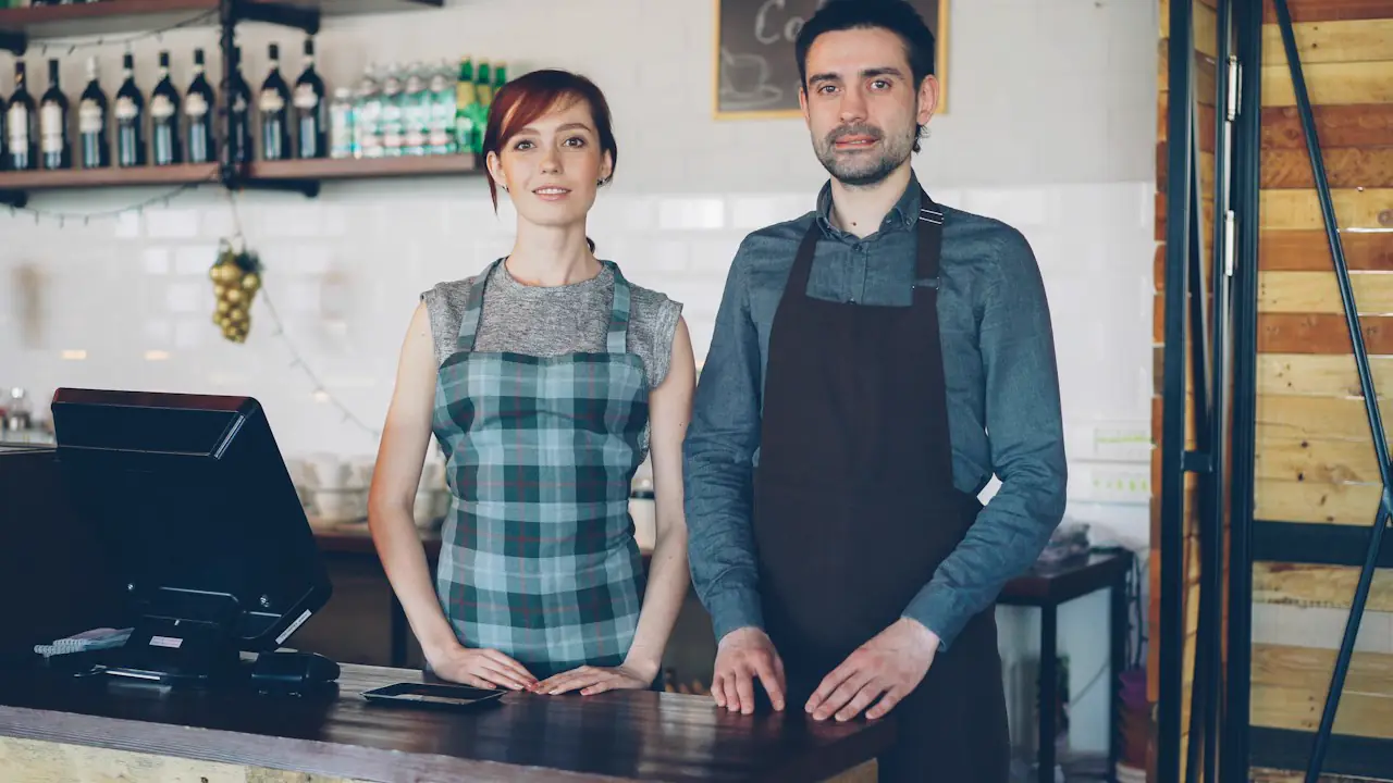 Two smiling cafe owners in aprons standing behind their counter