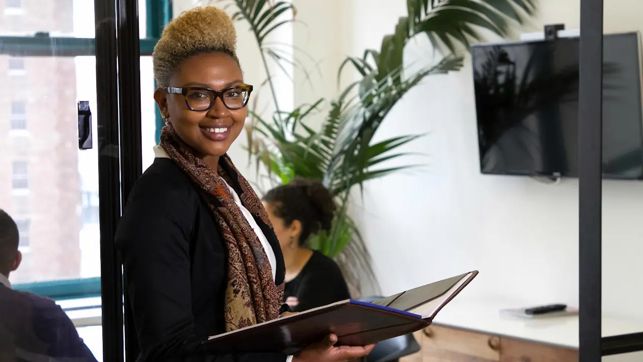 A smiling woman in glasses holding an open binder in a bright office