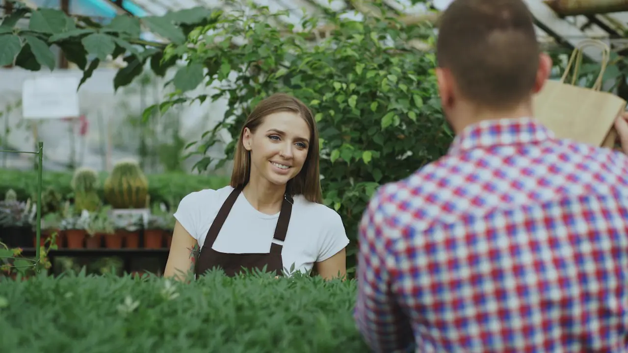 A smiling garden centre worker in an apron serving a customer among the plants