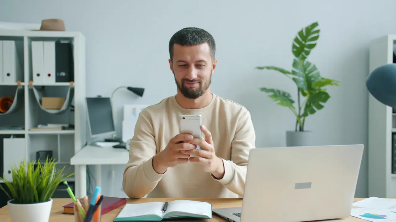 A smiling man checking his phone at a desk with a laptop