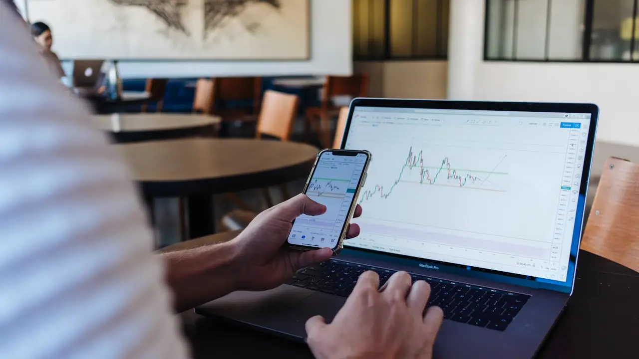 Hands holding a phone with a price chart in front of a laptop showing another chart