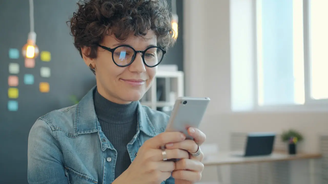 A smiling woman in glasses reading a message on her phone