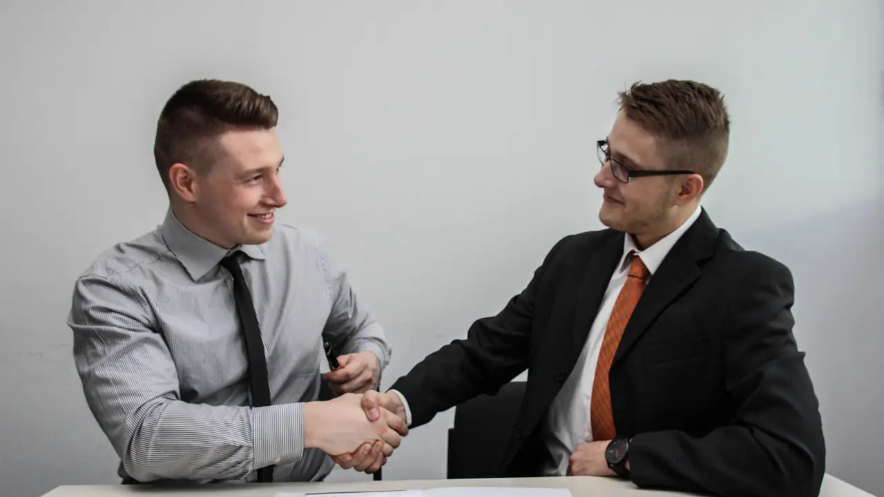 Two smiling men in shirts and ties shaking hands across a desk with papers on it