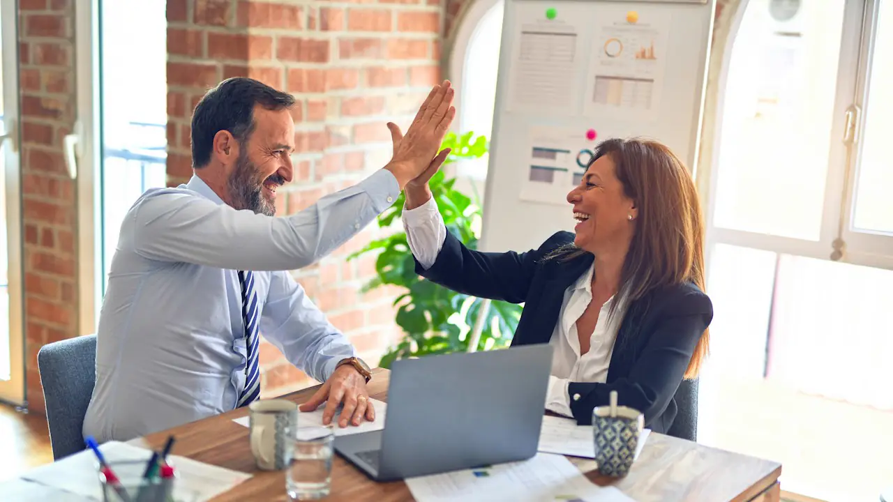 Two colleagues sharing a high five across a desk with a laptop and papers