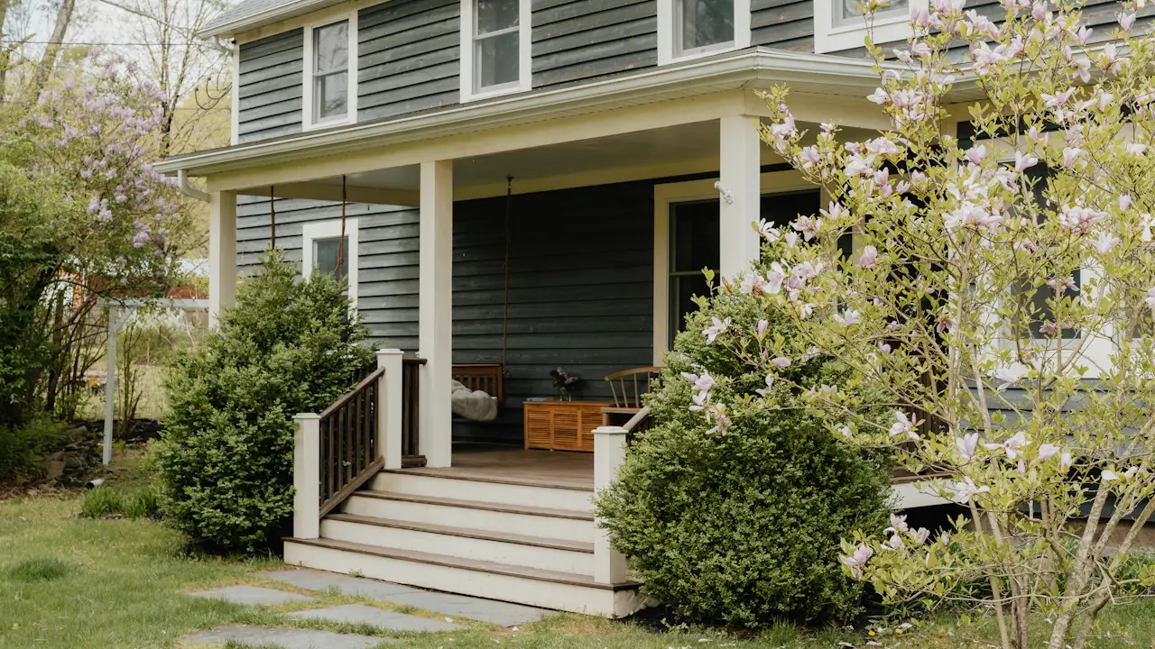 A grey two storey house with a wide front porch and a flowering tree