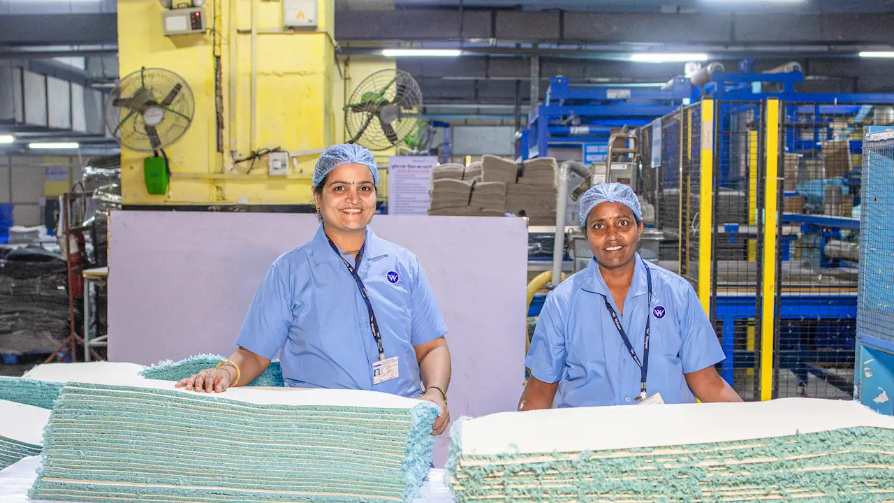Two smiling factory workers in blue coats and hairnets beside stacks of material
