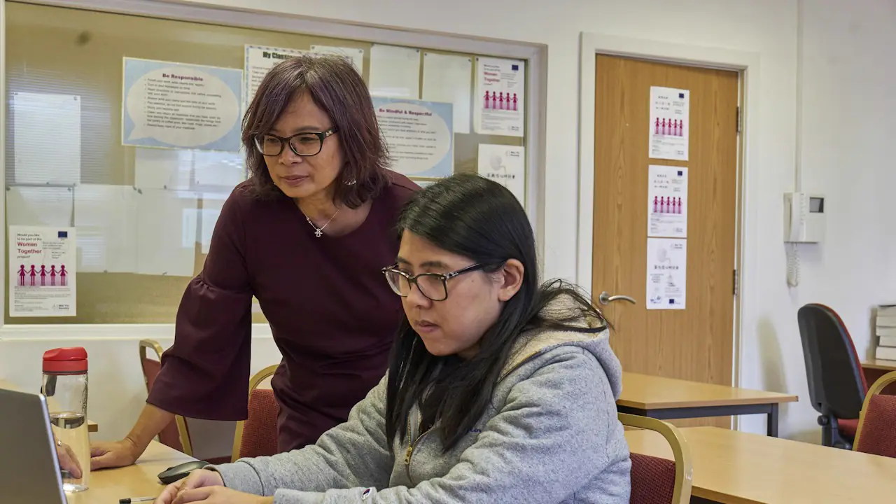 A tutor leaning in to help a student working at a laptop in a classroom