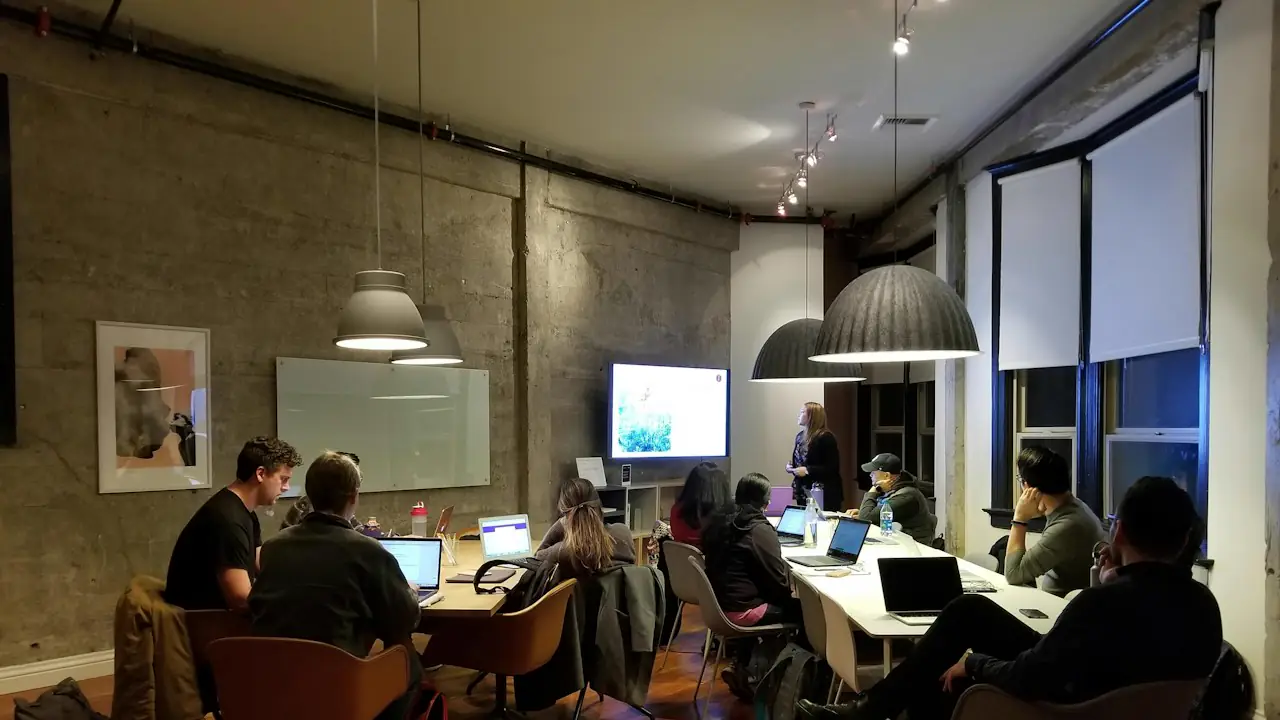 People working at laptops around long tables in a busy shared workroom
