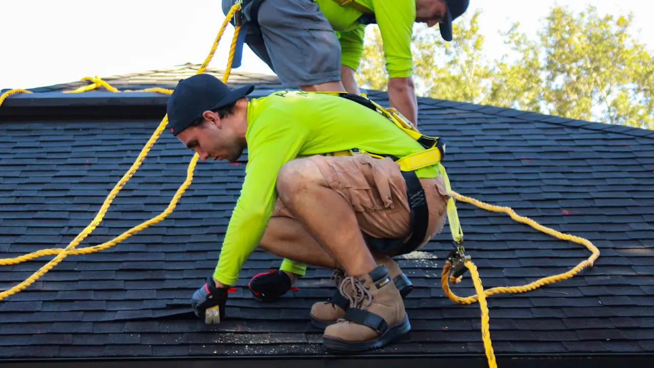 Two roofers in bright safety shirts and harnesses working on a shingled roof