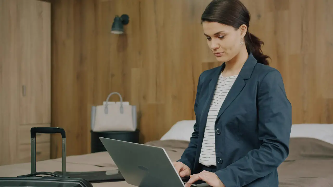 A woman in a blazer working on a laptop in a hotel room beside her suitcase