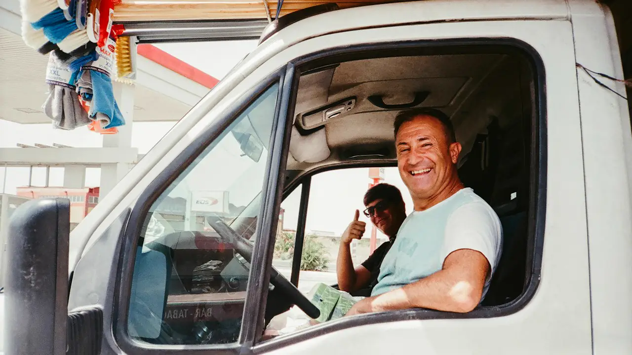 A smiling man at the wheel of a white work van, with a colleague giving a thumbs up