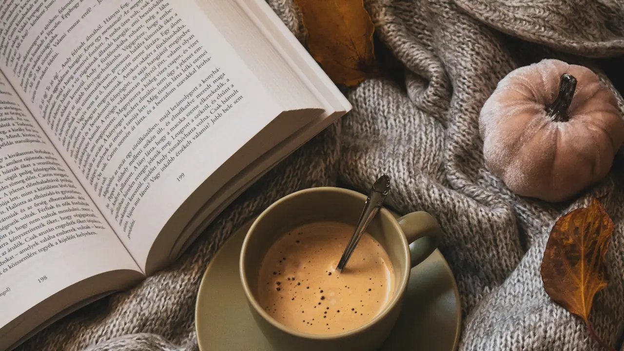 A cup of coffee and an open book on a knitted blanket with autumn leaves